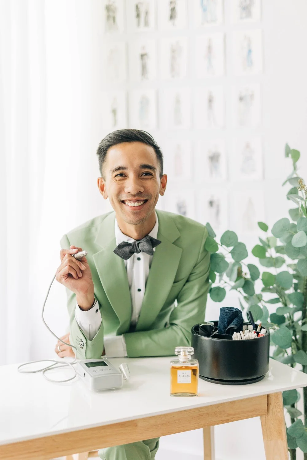 A live wedding artist in a sage suit sits at a clean workstation with a perfume bottle and tools, smiling at camera.