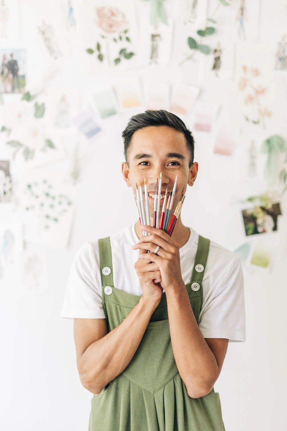 Smiling artist holds a fan of paintbrushes in front of a wall covered with watercolor sketches and color swatches, giving playful, creative studio energy.