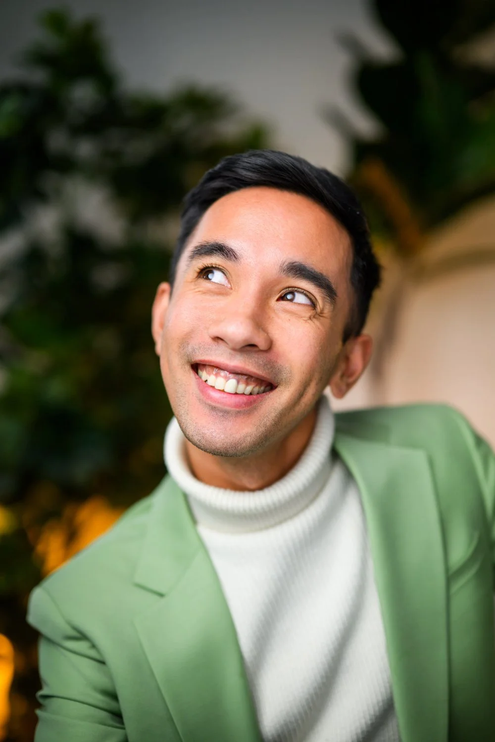Smiling person in a sage green blazer and cream turtleneck looks up and to the side, with soft bokeh greenery behind—warm, approachable portrait with modern, polished energy.