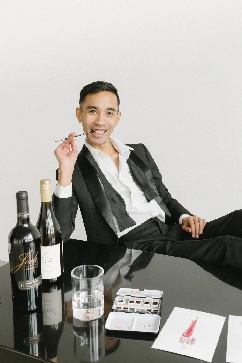 Artist in a tux sits at a glossy table with wine bottles, water, and paint set, posing with a brush—fashion-forward scene for wine country weddings and live winecolor guest portraits.