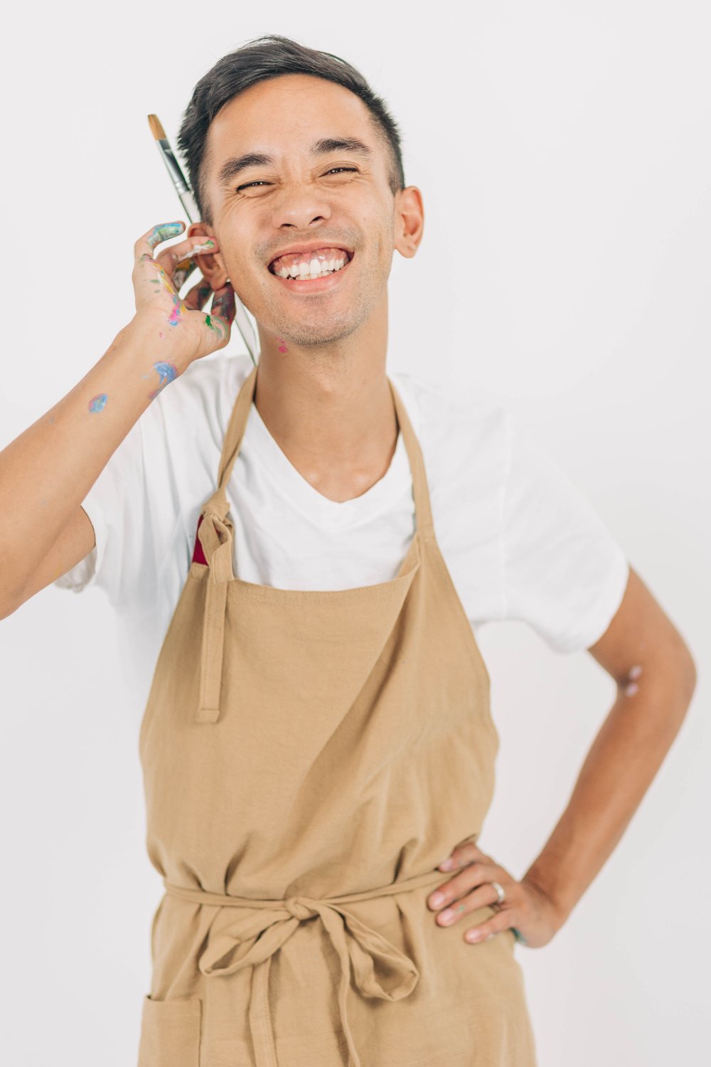 Close-up of an artist smiling in a tan apron, paint on his hand and a brush tucked by his face—behind-the-scenes artist portrait for an eco-friendly wedding blog, showing the hands-on, joyful process of making live keepsakes.