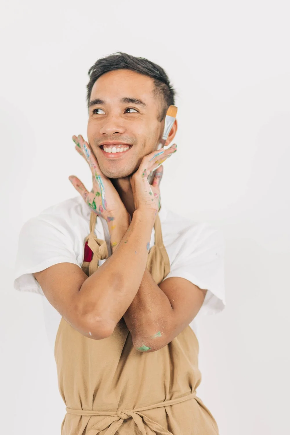 Playful studio portrait of a smiling artist in an apron in front of a bright white background. Paint-splattered hands framing his face while holding a paint brush.