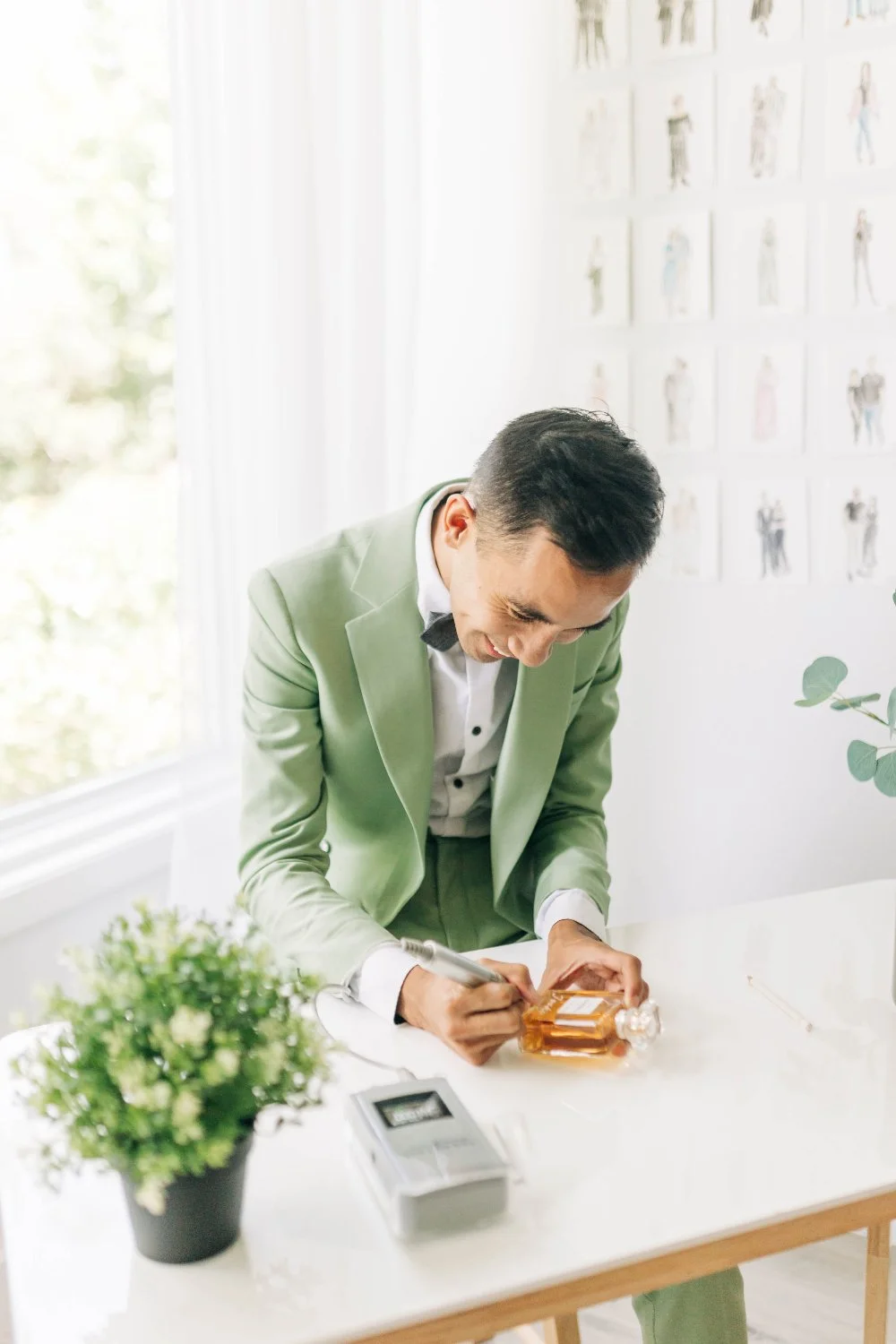 Artist in a sage-green suit engraves a perfume bottle at a bright white table, creating a personalized keepsake for guests. Clean studio light, greenery accents, and modern tools capture the vibe of luxury wedding entertainment ideas in action.