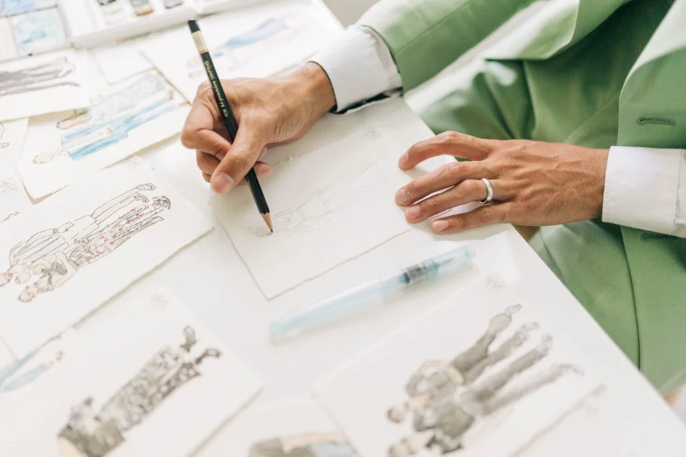 Close-up of hands sketching a fashion portrait on textured paper, surrounded by finished mini portraits.