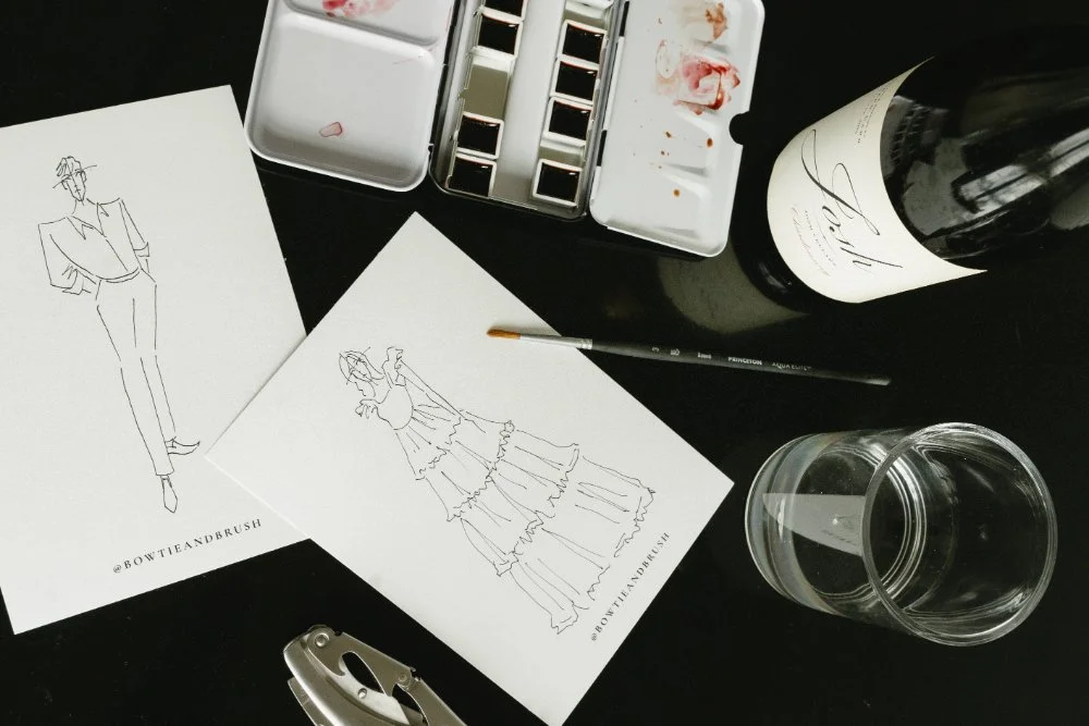 Flat lay of fashion sketches, watercolor palette, brush, wine bottle, and glass on a dark tabletop—tools and mood that inspire wine country weddings and on-site winecolor guest portraits.