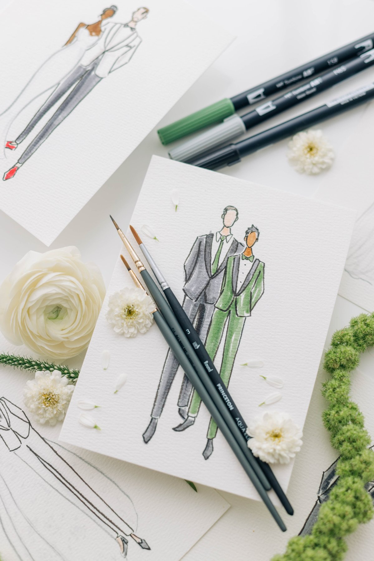 Flat lay of watercolor fashion portraits with paintbrushes, pens, and white blooms on textured paper—an elevated sustainable wedding favor idea that feels intentional, reusable, and frame-worthy instead of disposable.