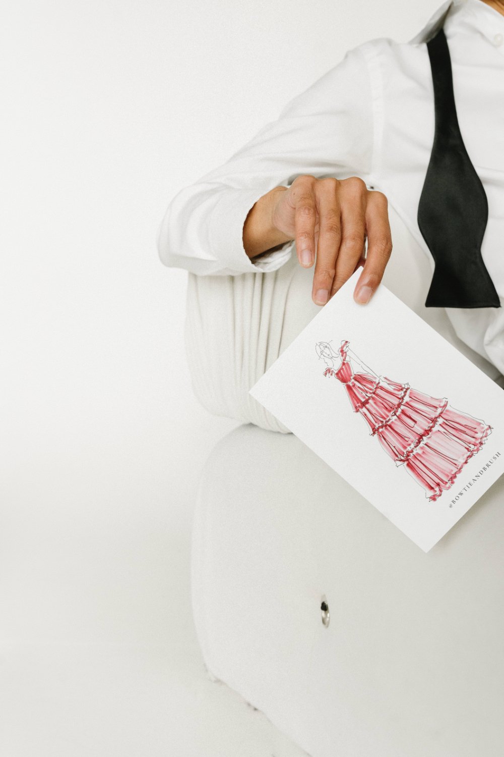 Hand in a crisp white shirt and black bow tie holds a fashion sketch with a red dress—editorial detail for wine country weddings, showing the chic vibe behind winecolor guest portraits.