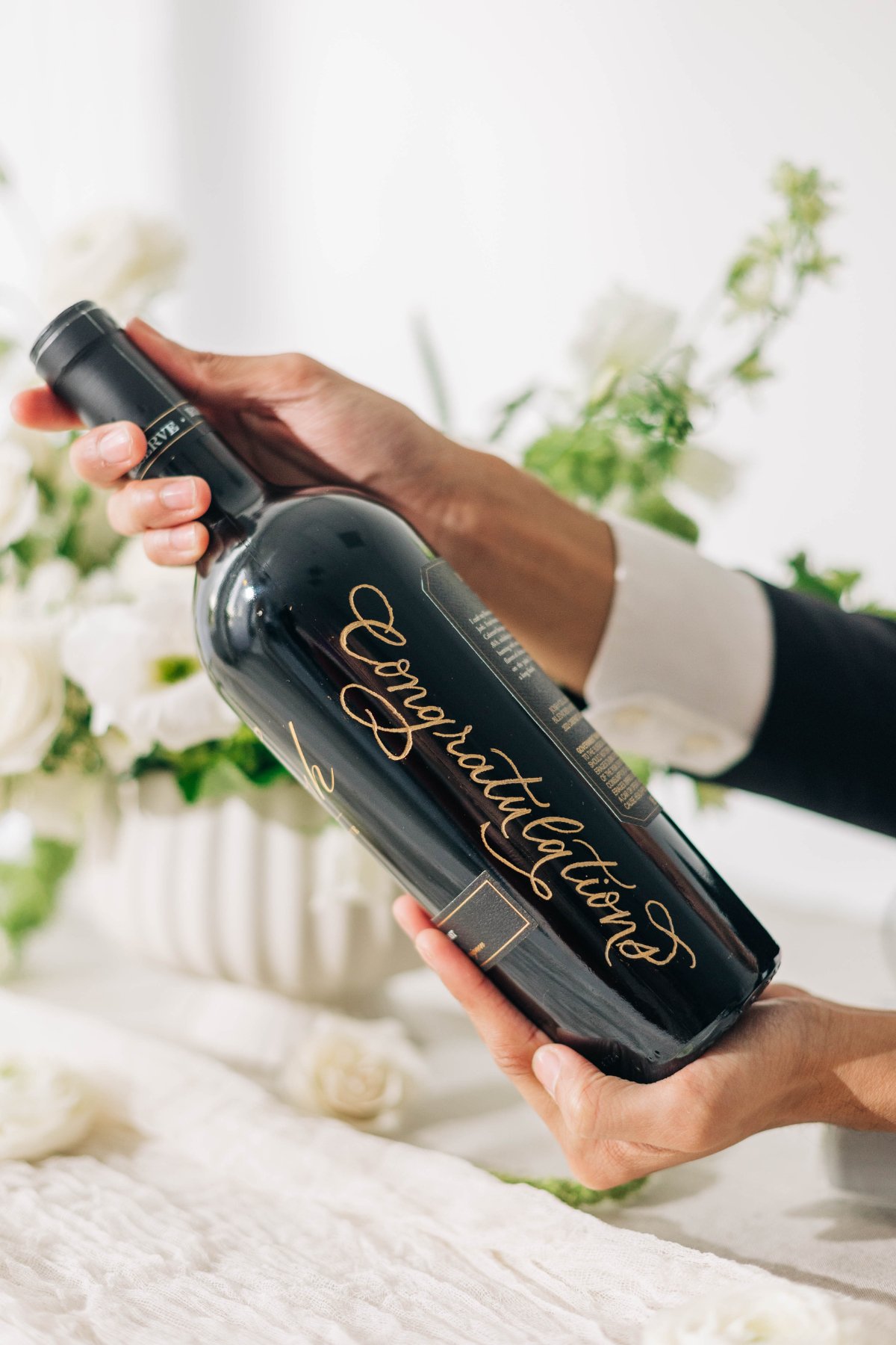 Close-up of hands presenting a wine bottle with elegant gold calligraphy lettering, styled with white florals for a luxe reception detail.