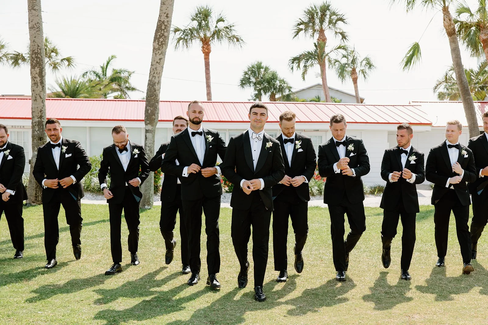 Groom and groomsmen in black tuxedos walking on grass at outdoor wedding ceremony, with palm trees and a building with a red roof in the background.