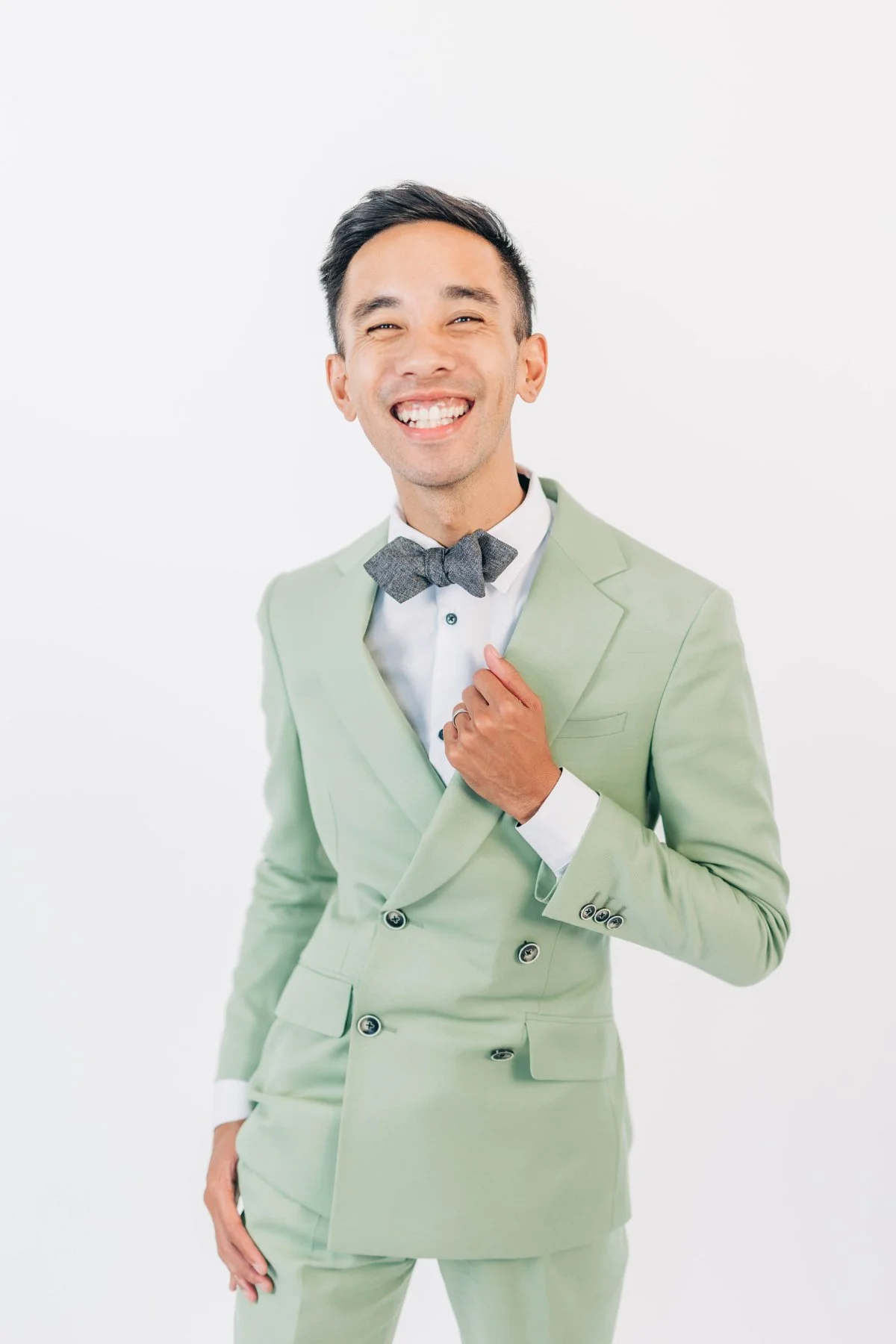 Bright studio portrait of a cheerful man in a mint green suit and gray bow tie, adjusting his lapel on a clean white background. Polished and playful branding that pairs perfectly with modern alternatives to traditional wedding favors.