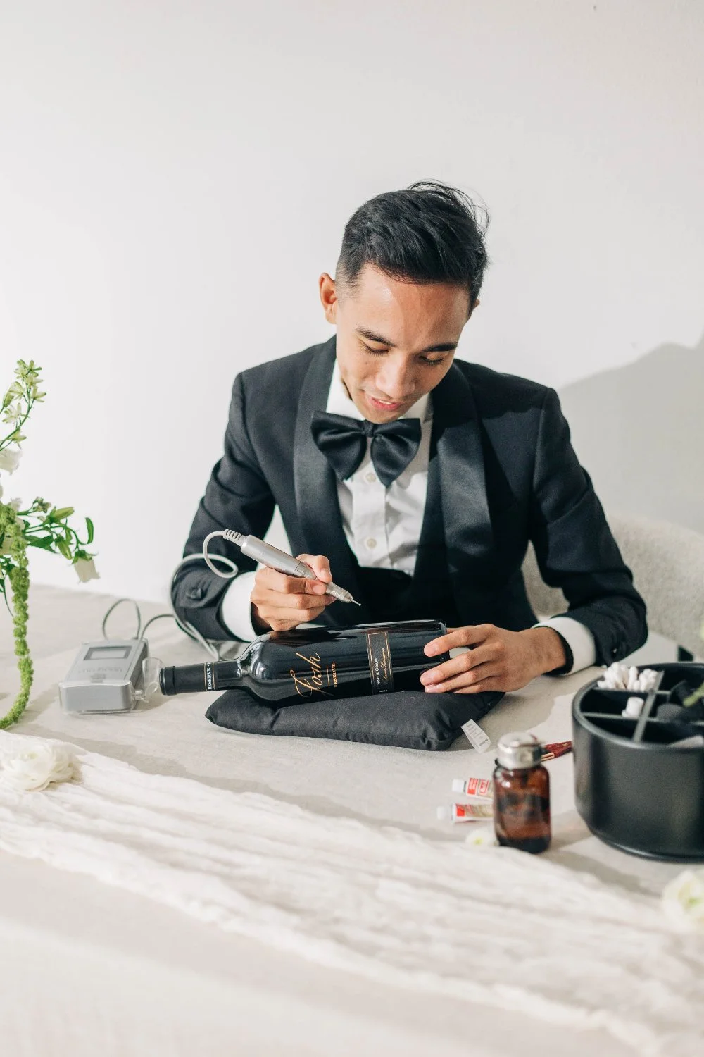 Dressed in a black tux and bow tie, the artist engraves a wine bottle resting on a cushion, surrounded by tools and floral greenery. The scene feels sleek and intimate—an on-the-spot personalization moment guests love.