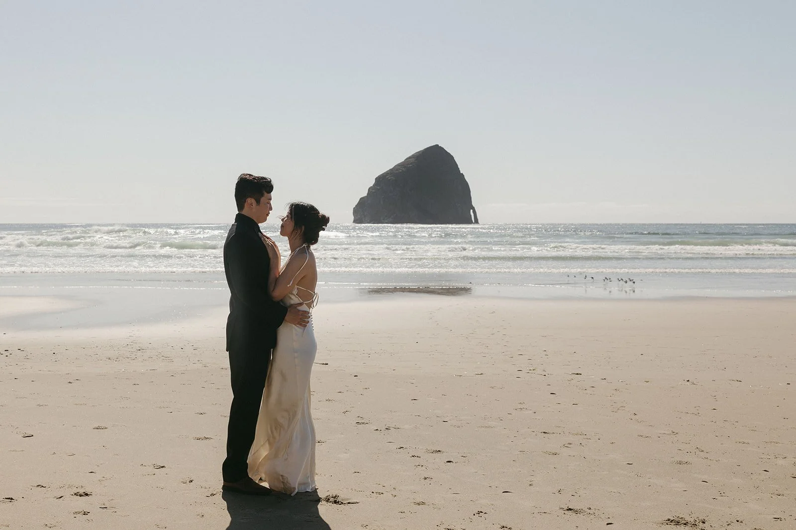 A man and woman in wedding attire standing close on a beach, with the ocean and a large rock formation in the background.