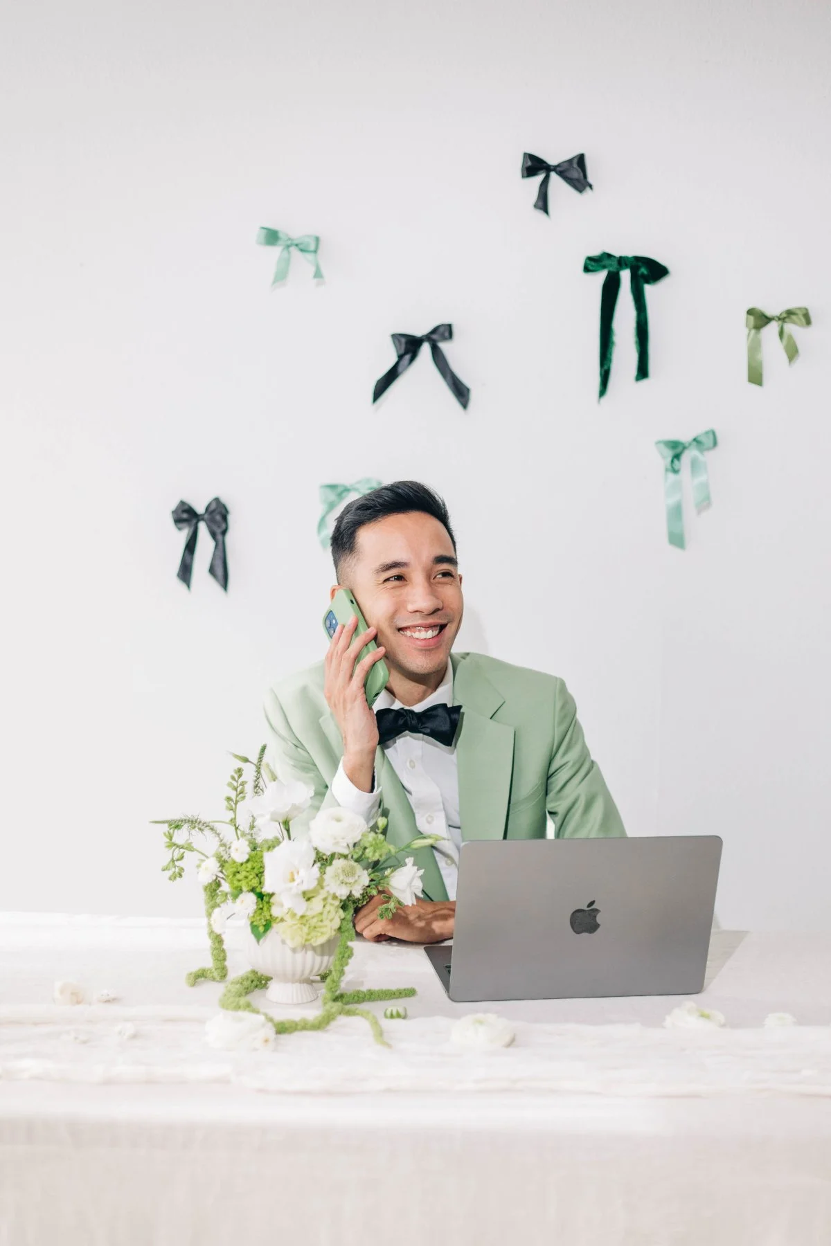 Smiling artist in a mint suit and bow tie sits at a styled desk with laptop and lush white-green florals, holding a phone. Ribbon bows decorate the wall.