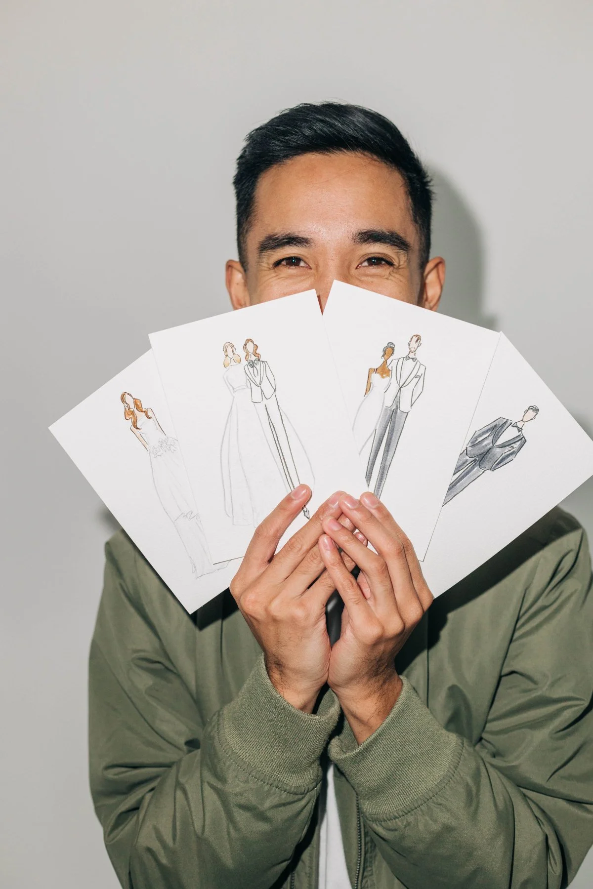 Close-up of a smiling man holding a fan of watercolor fashion portrait cards in front of his face against a gray wall. The hand-painted couples and outfits hint at live art keepsakes that feel more personal than traditional wedding favors.