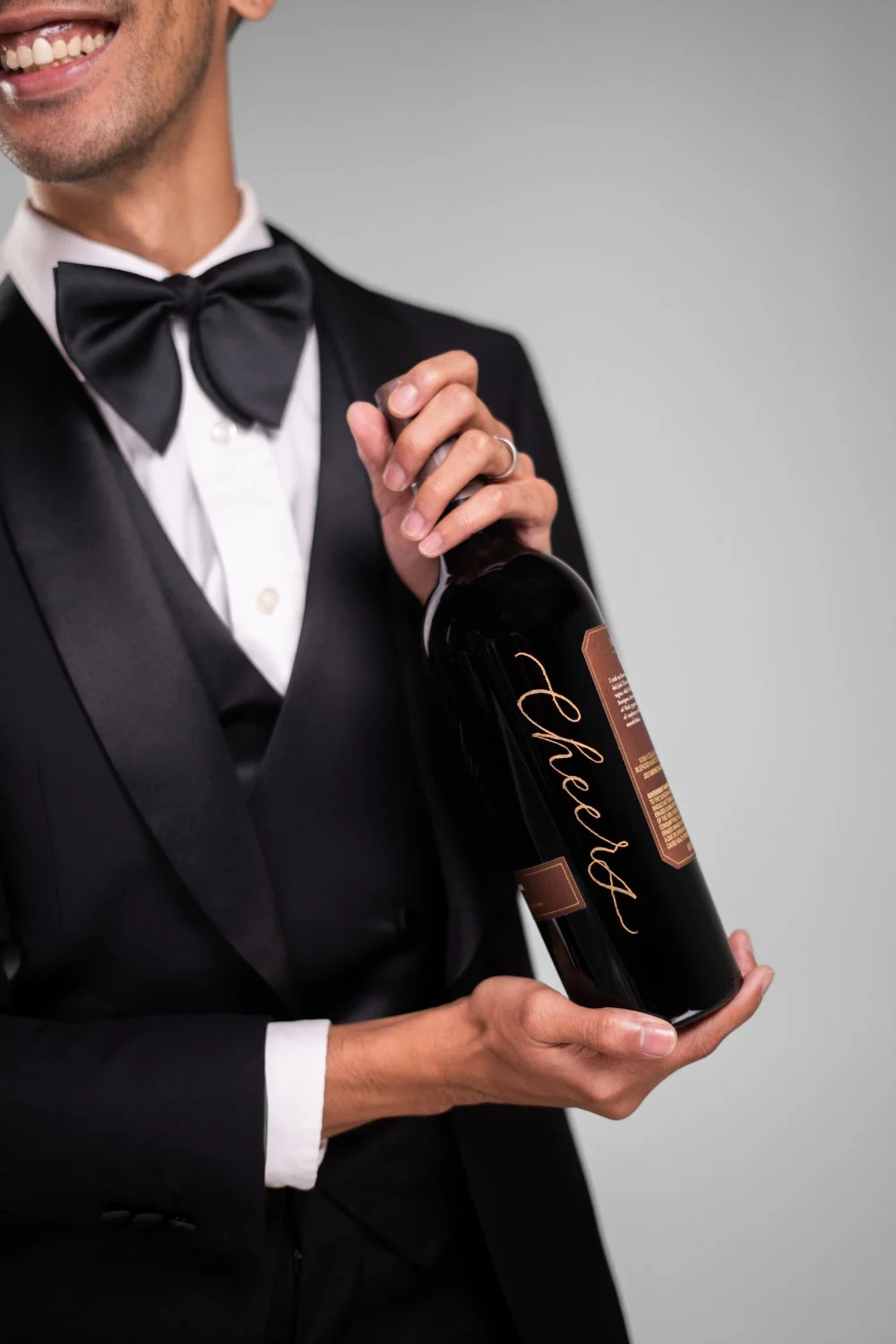Close-up of a tuxedo with an oversized black bow tie holding a dark bottle with elegant script lettering—sleek, celebratory detail shot.