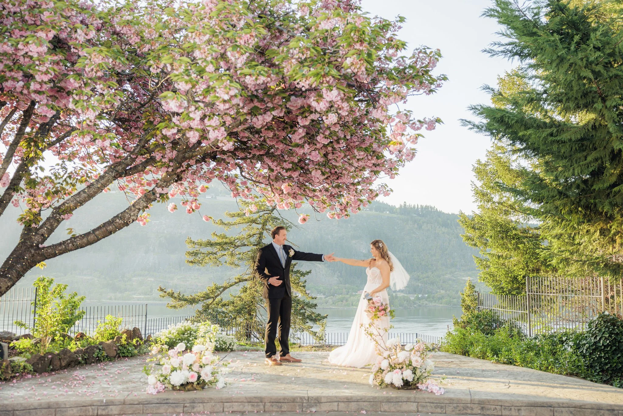 A bride and groom holding hands during their wedding ceremony outdoors, with pink cherry blossoms overhead and a scenic lake and mountain background.