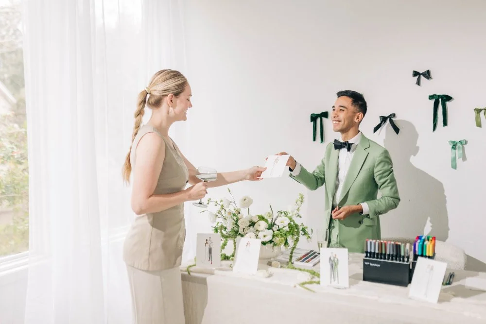 Live artist in a sage suit hands a guest her finished fashion-style portrait at a bright event table with florals and markers—an interactive keepsake moment perfect for a wedding or brand activation.