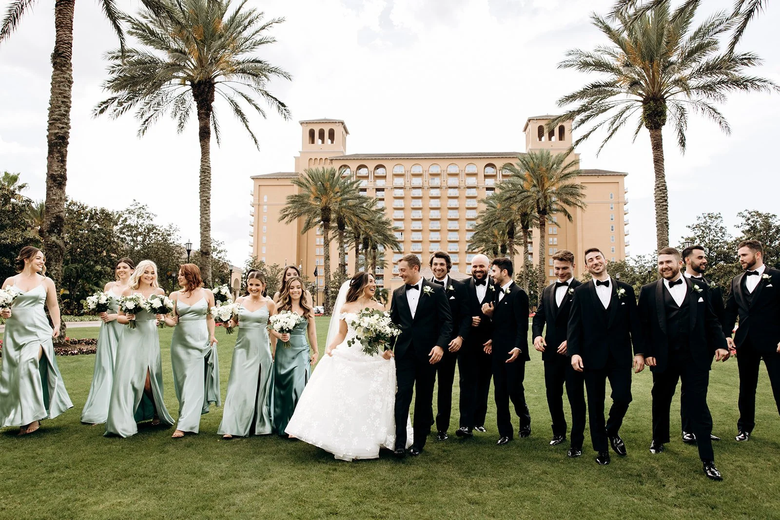 A wedding party with bridesmaids in light green dresses and groomsmen in black tuxedos, walking on a lawn with palm trees and a large hotel or resort building in the background.