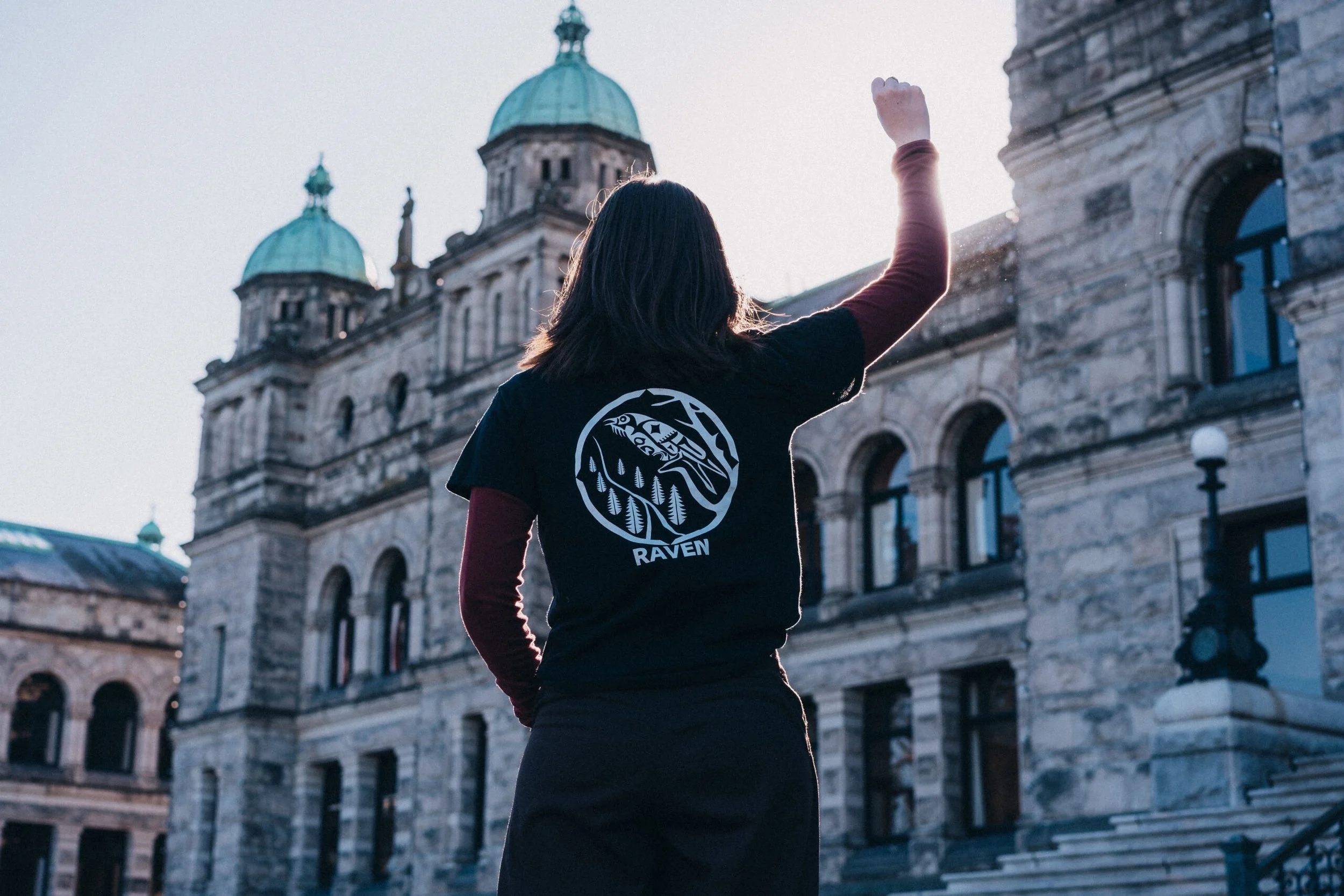 Woman wearing a Raven logo t-shirt raising her right hand in front of a building