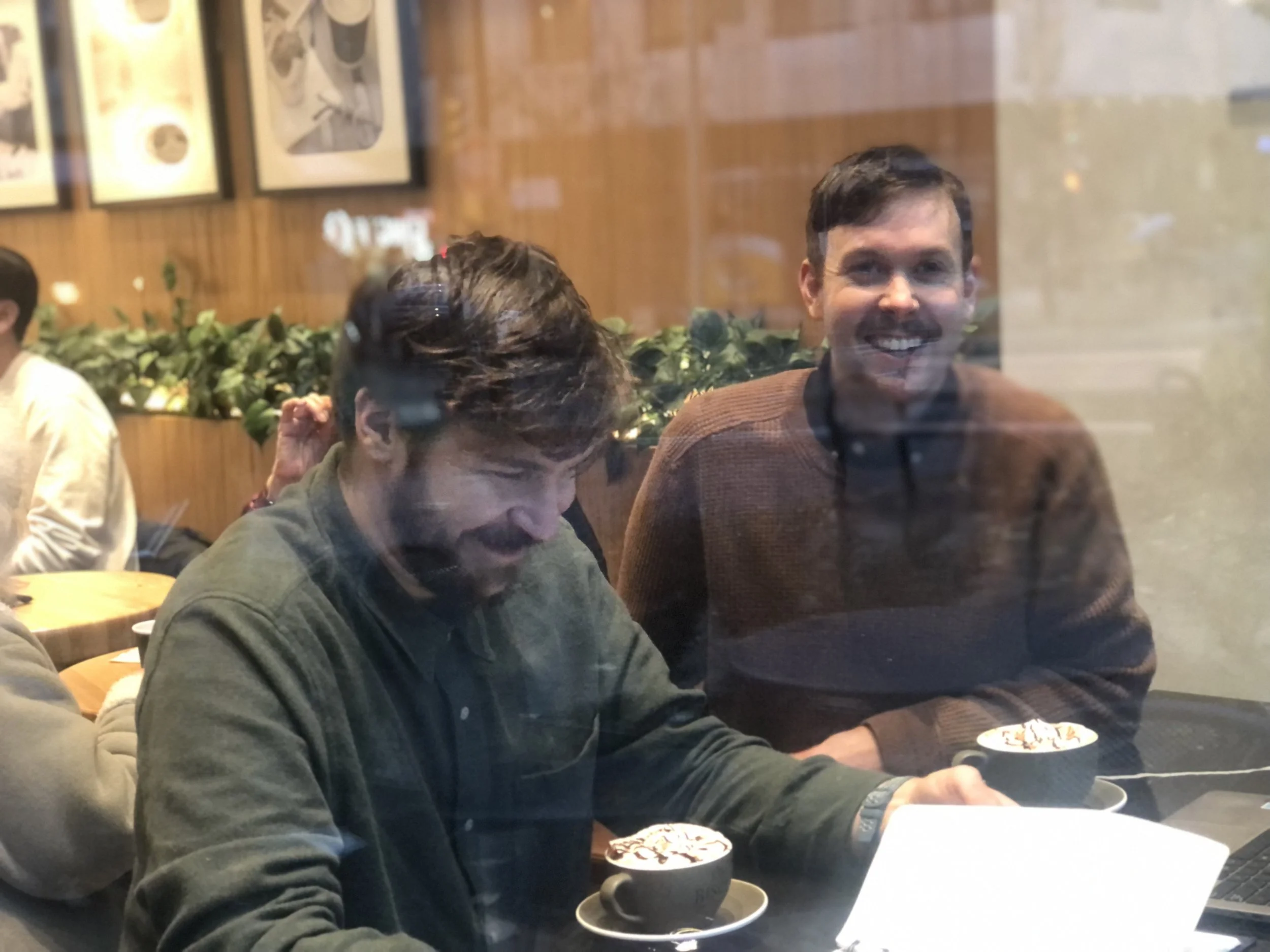 Two men sitting at a table in a cafe, seen through a window. One man is smiling, and the other is looking down at a laptop with drinks in front of them.