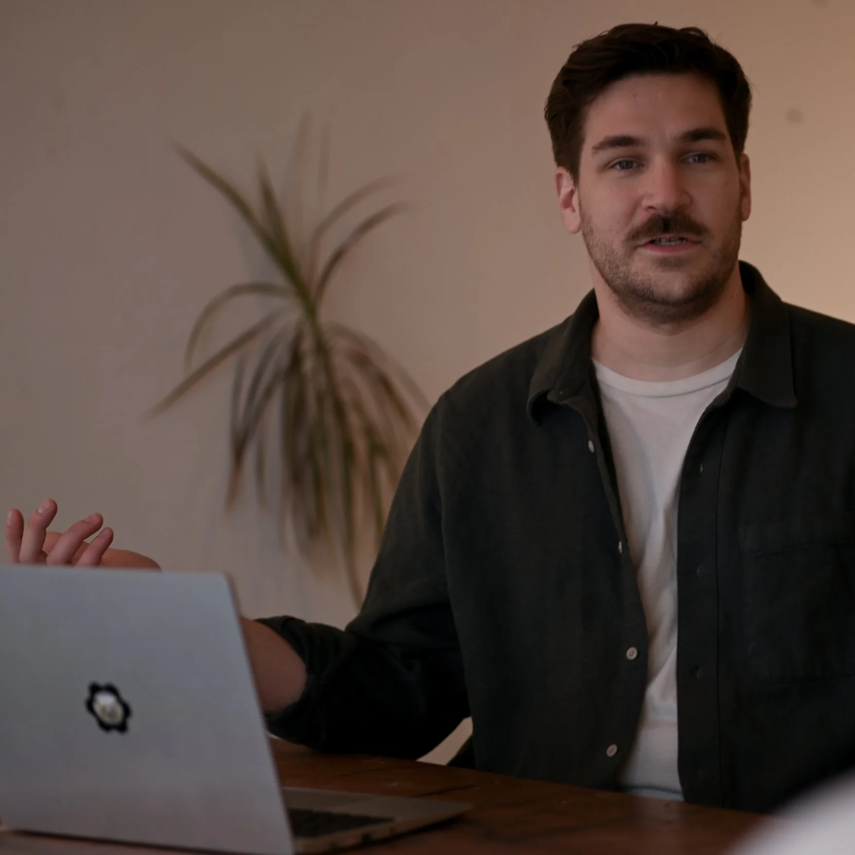 Man at a table with a laptop, gesturing with his hand