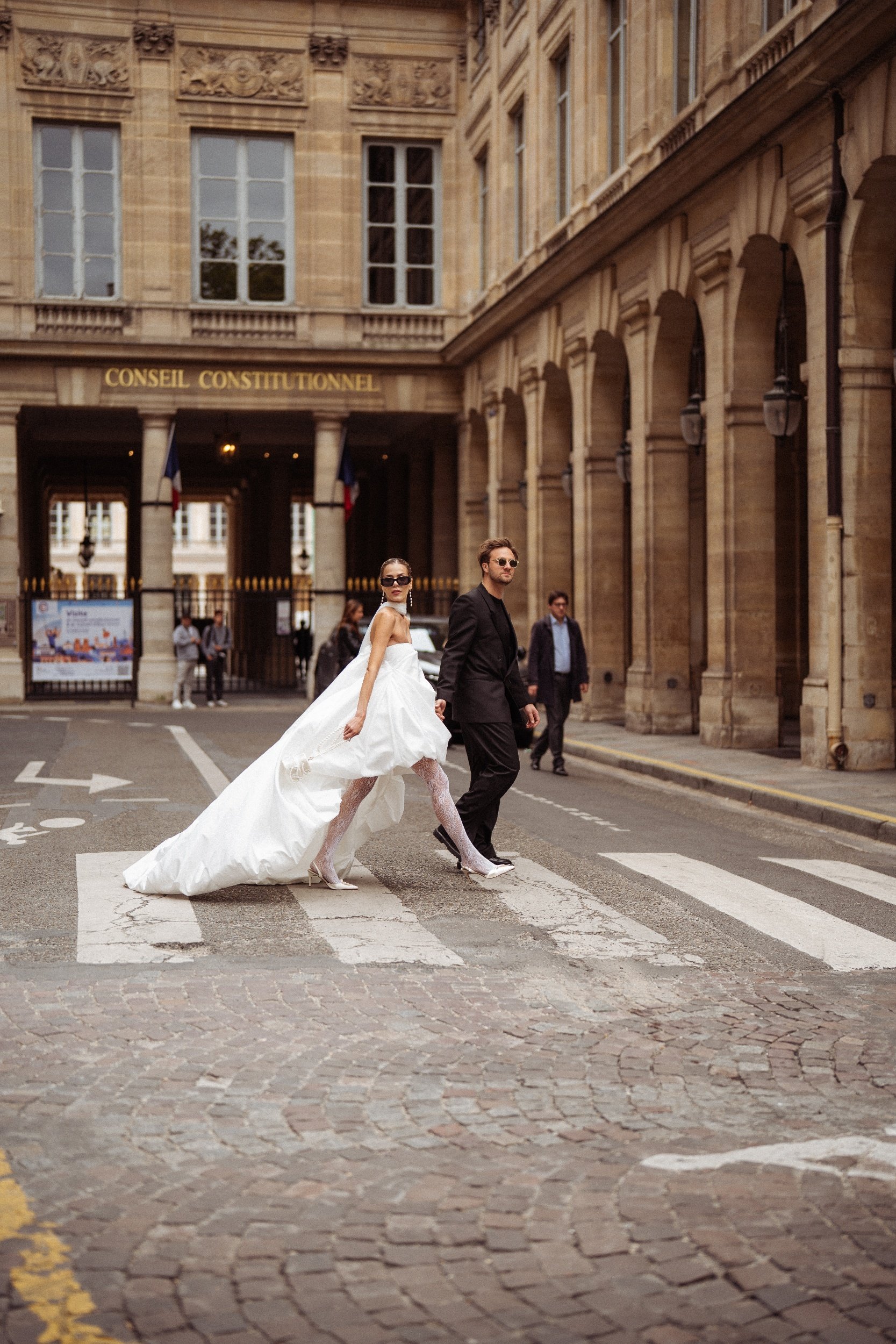 man in suit and woman in wedding dress walking across a parisien street