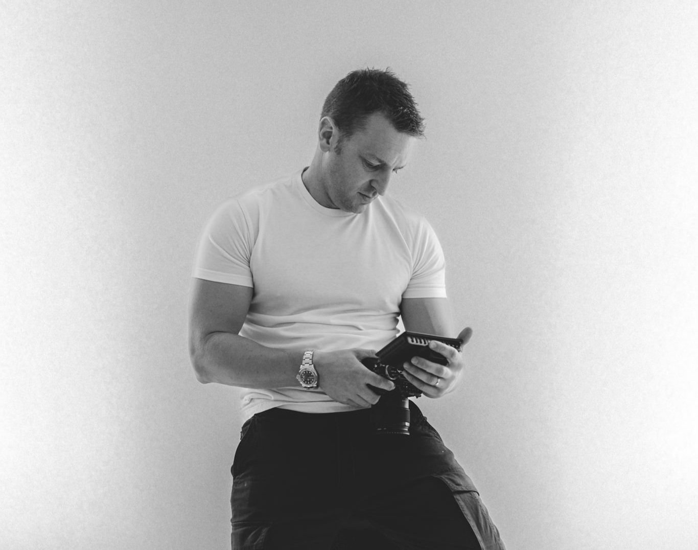 man holding a video camera sat on a stool, looking at the camera monitor