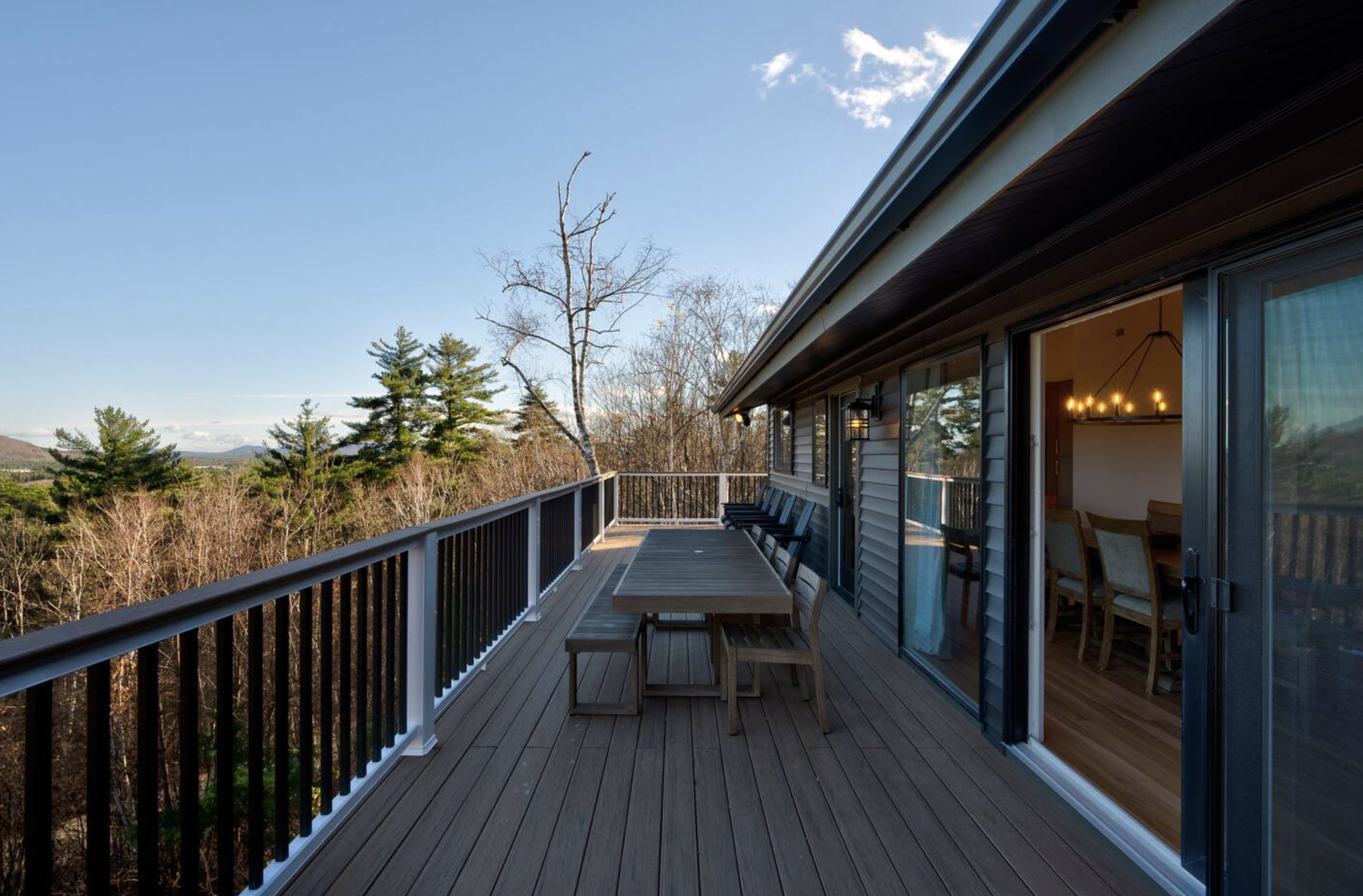 Balcony with wooden flooring, black railing, outdoor table, and chairs overlooking trees and distant mountains, with a blue sky and some clouds.