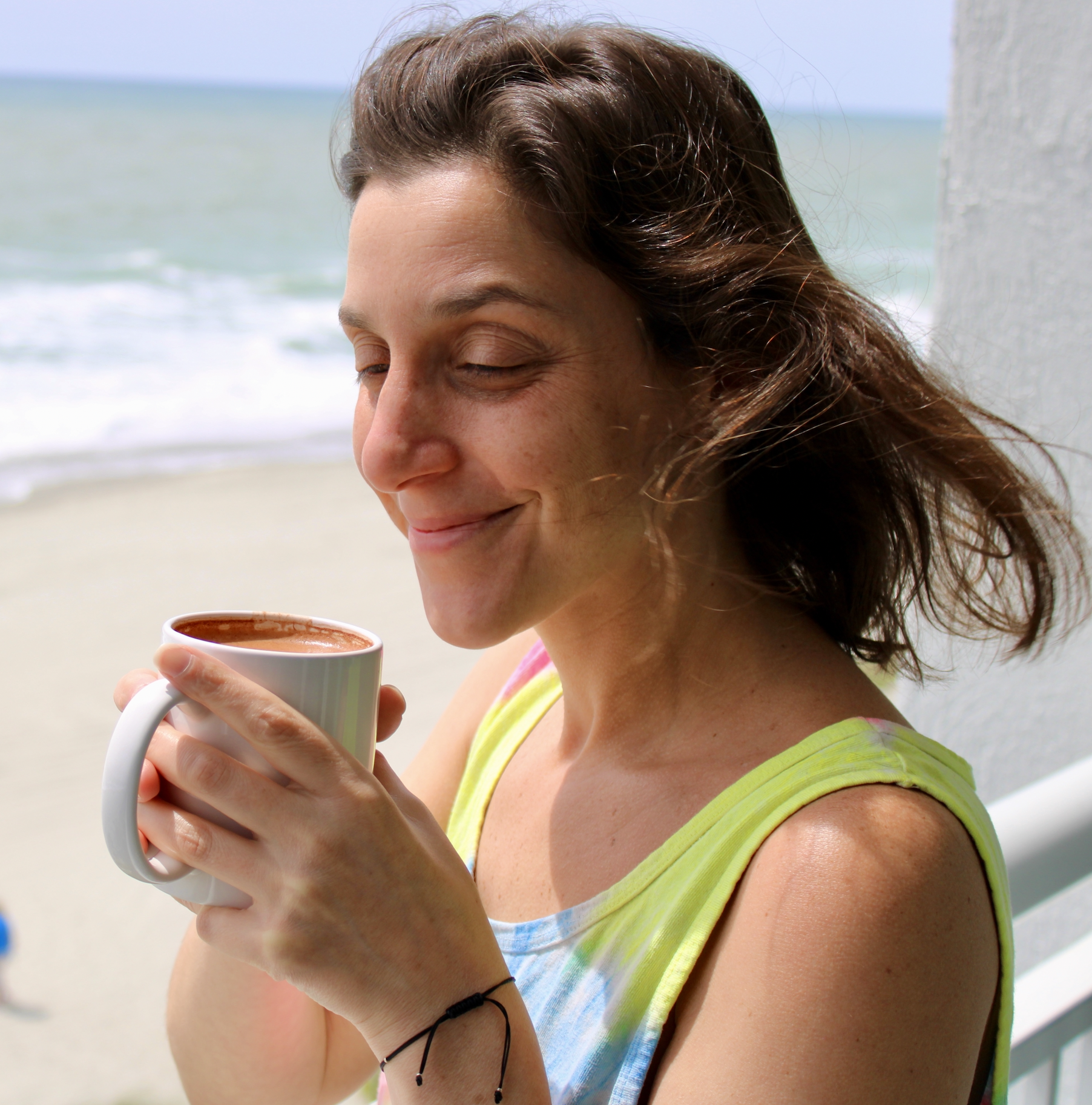 A woman with wavy brown hair smiling with her eyes closed, holding a white mug with a hot beverage, standing outdoors at the beach with sand and ocean in the background.