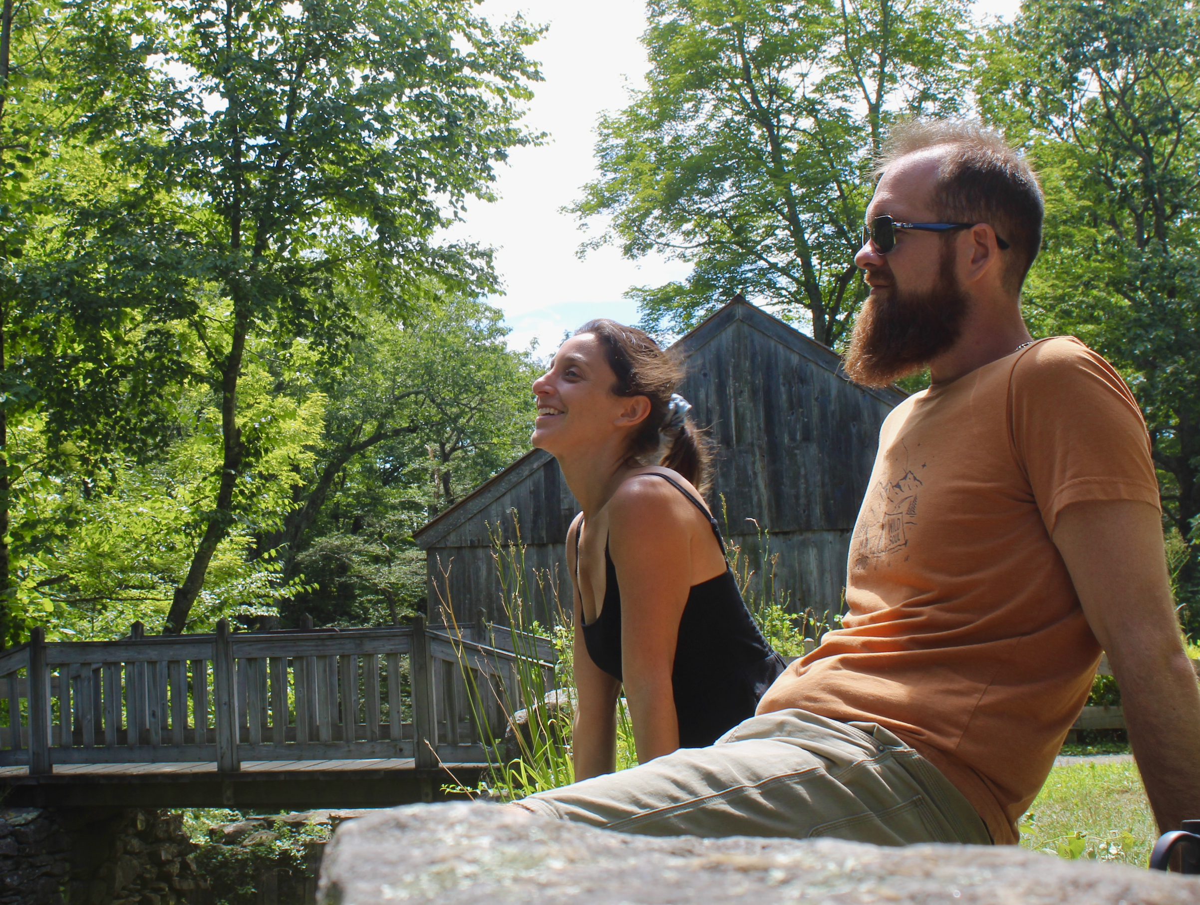 Two people smiling, sitting outdoors on a sunny day among green trees, with a wooden fence and shed in the background.