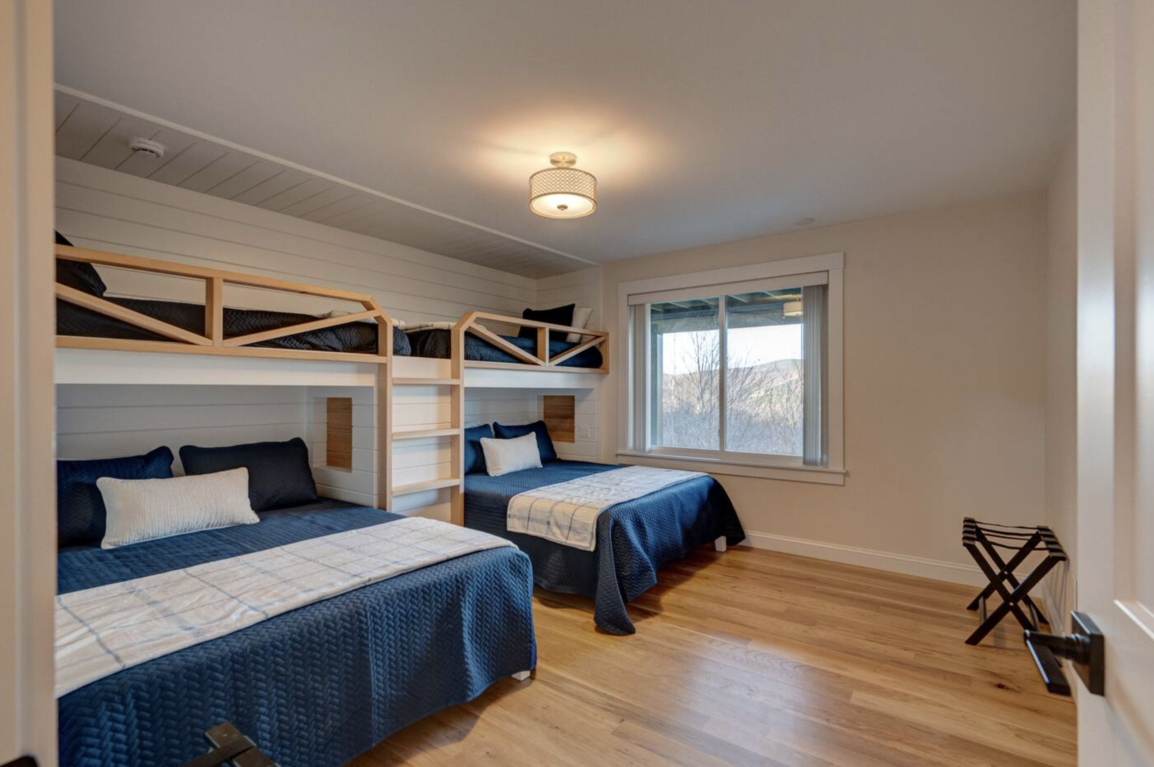 A bedroom with three twin beds, each with blue bedspreads and pillows, a window showing a view of trees and mountains, and a ceiling light. There is a luggage rack and folding luggage racks in the room.