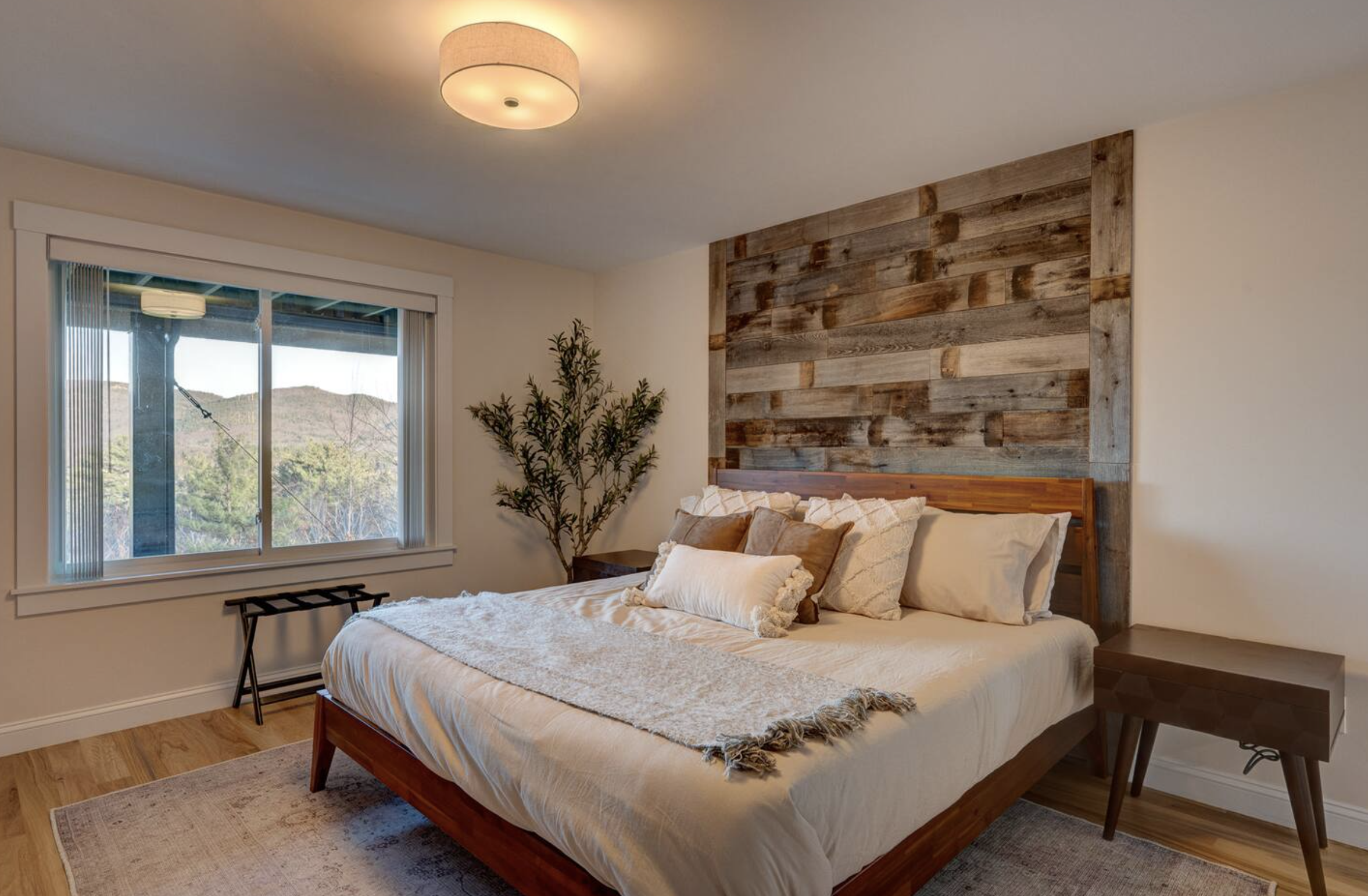 A bedroom with a wooden headboard, white bedding and pillows, a potted plant, a window with blinds, and a ceiling light.