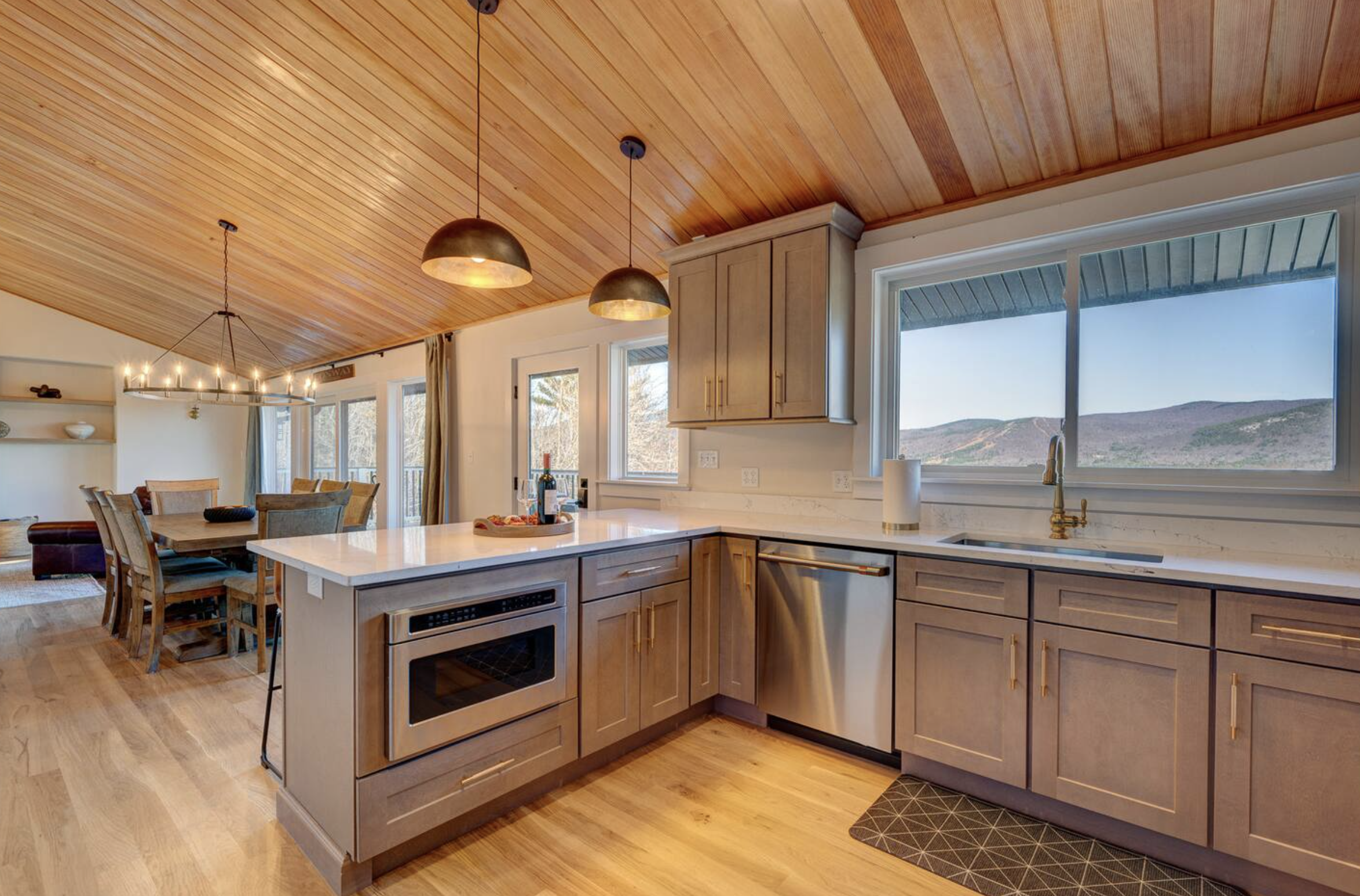 Kitchen with wooden cabinets, marble countertops, large windows with scenic mountain views, modern pendant lights, and a dining area in the background.