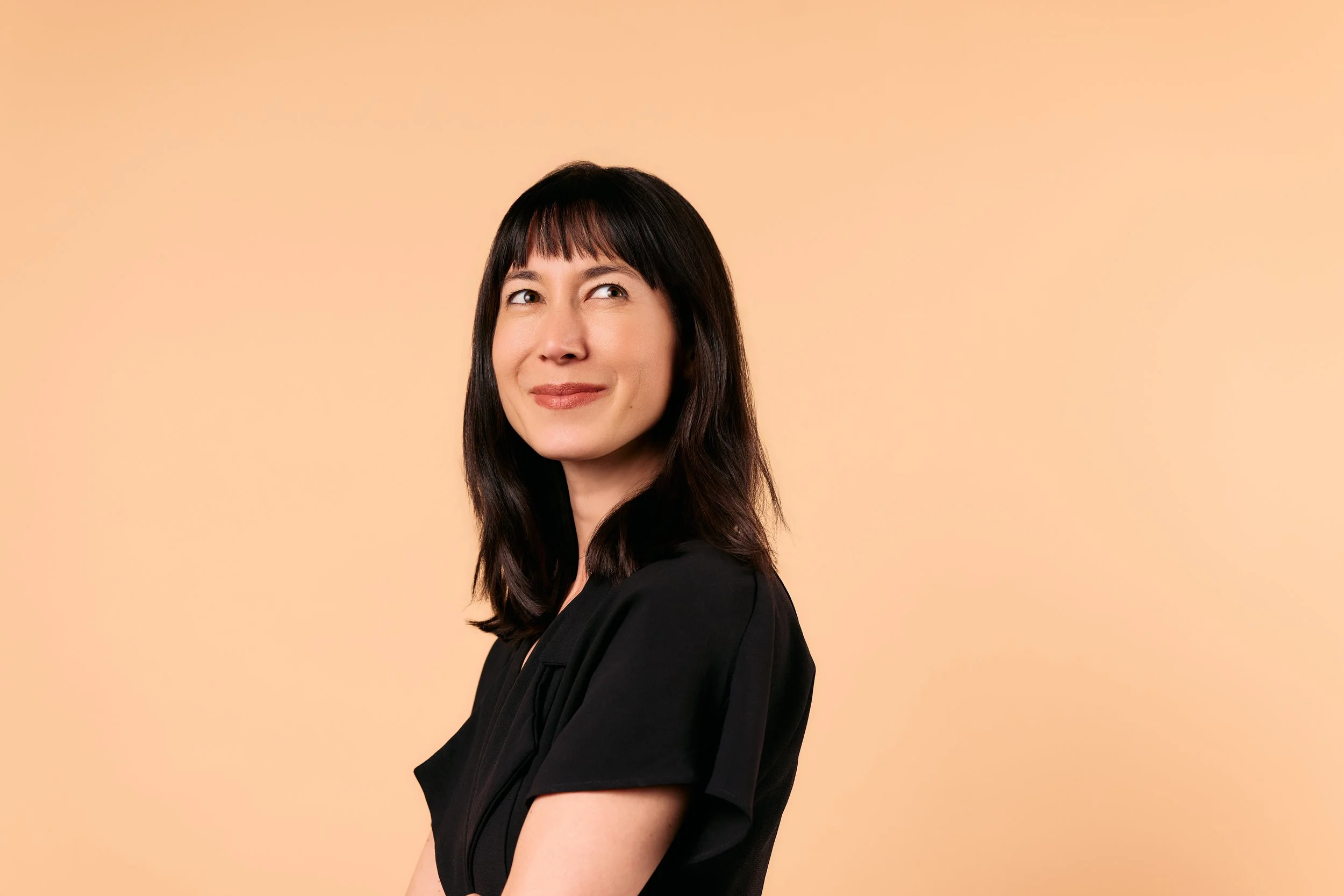 A woman with dark hair and bangs, wearing a black top, smiling with her head slightly tilted to the side against a plain peach background.