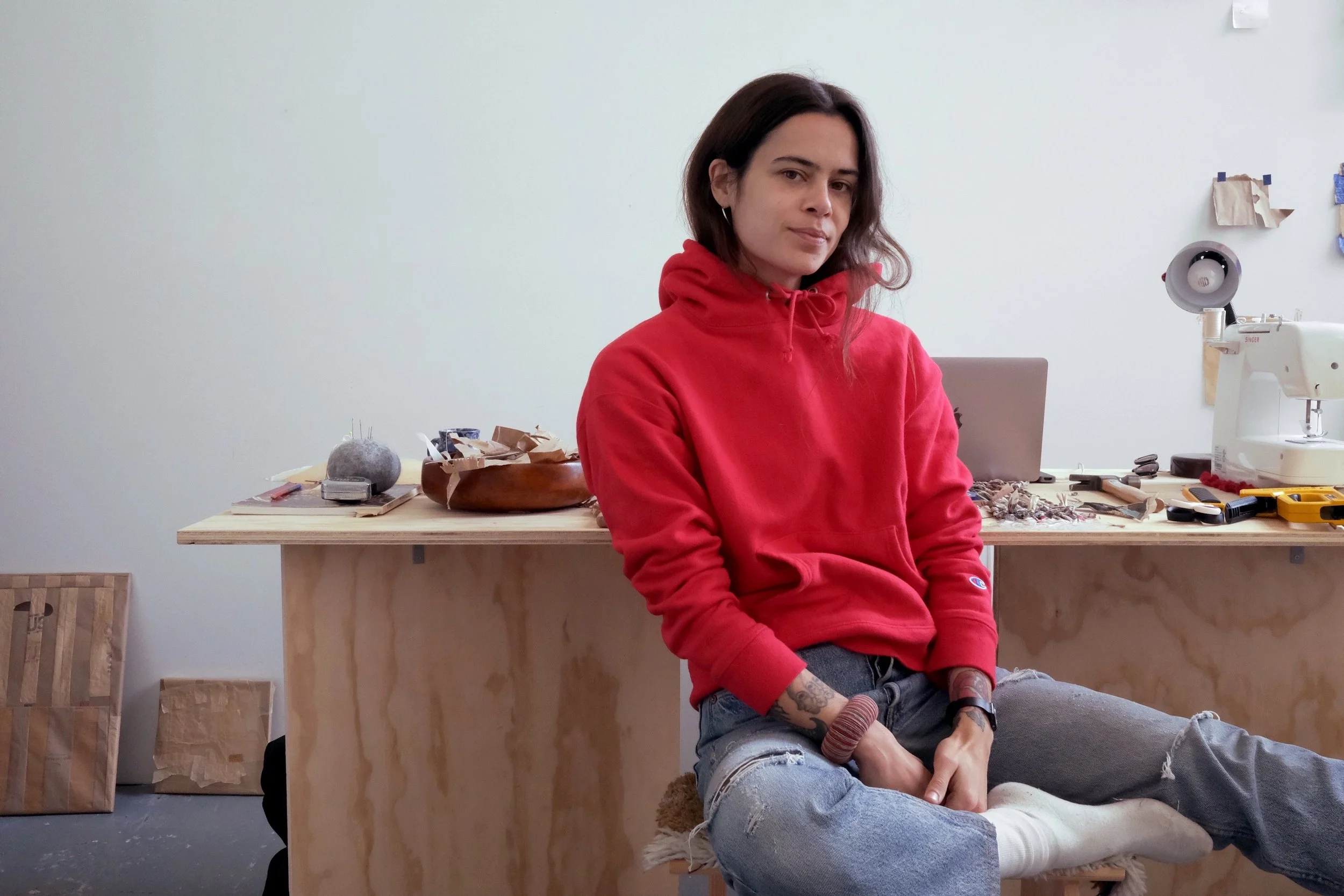A woman with dark hair sitting on a stool in front of a workbench, wearing a red hoodie and ripped jeans, in a woodworking or crafting studio.