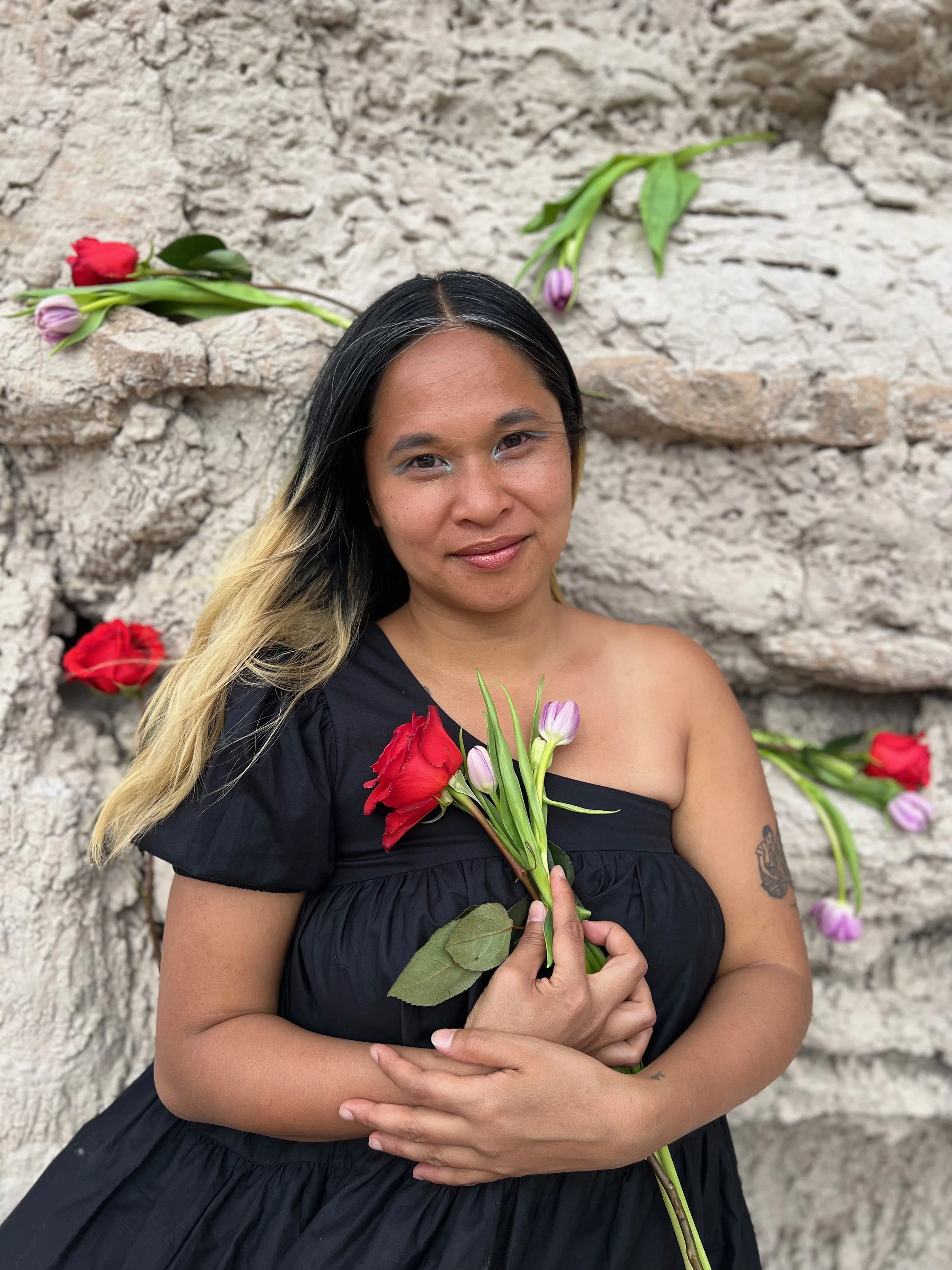 Woman with long black hair and blonde highlights holding a bouquet of red and purple flowers, standing in front of a stone wall with additional flowers placed on the wall.
