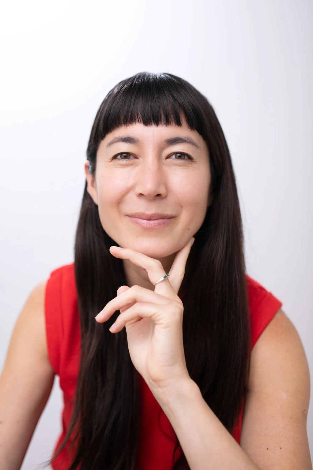 A woman with long dark hair and bangs, wearing a red sleeveless top, smiling softly while touching her chin with her hand against a plain white background.