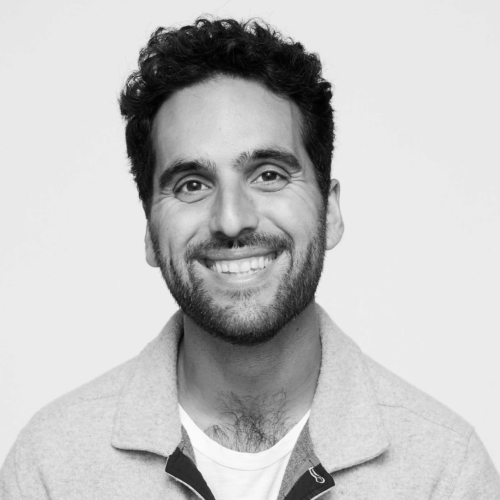Black and white portrait of a smiling man with curly hair and a beard, wearing a light-colored jacket over a T-shirt.