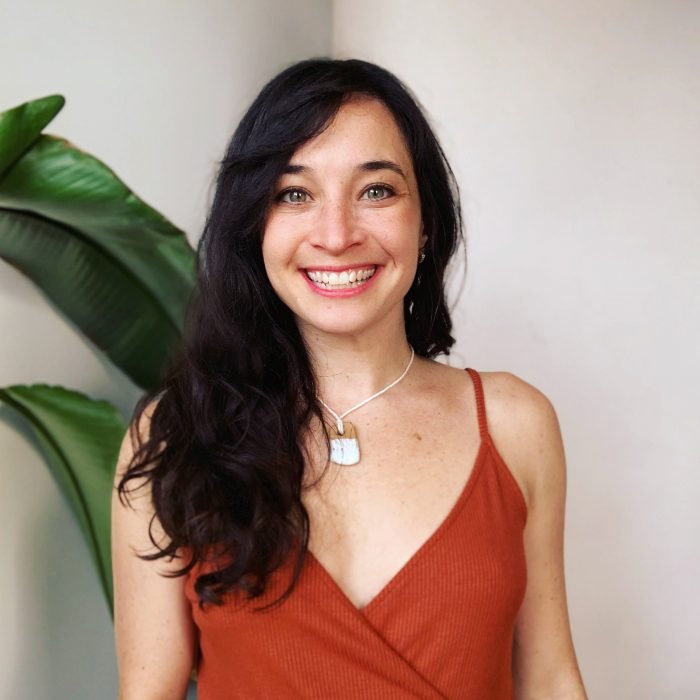 A woman with long wavy dark hair smiling in front of a plant and white wall, wearing a rust-colored spaghetti strap top and a necklace.