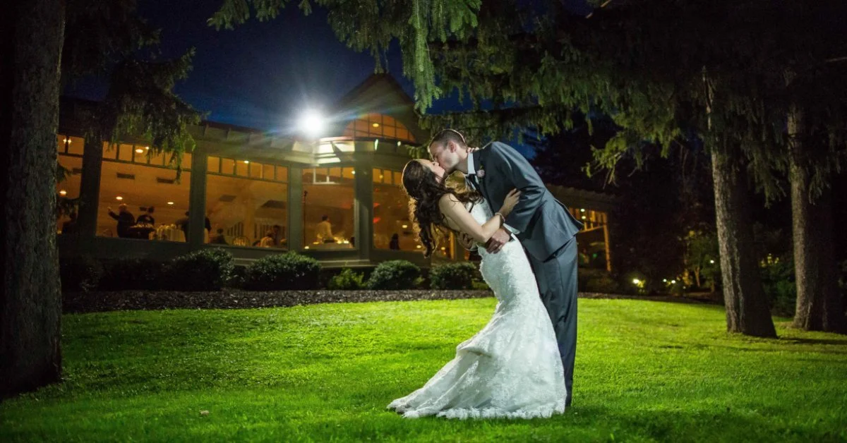 A bride and groom share a kiss on a lawn at night, illuminated by venue lighting, with a reception building glowing warmly in the background beneath tall evergreen trees.