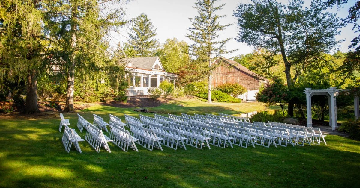 A garden wedding venue featuring neatly arranged white chairs on a grassy lawn, with a white arbor, wooded landscape, and rustic buildings in the background.