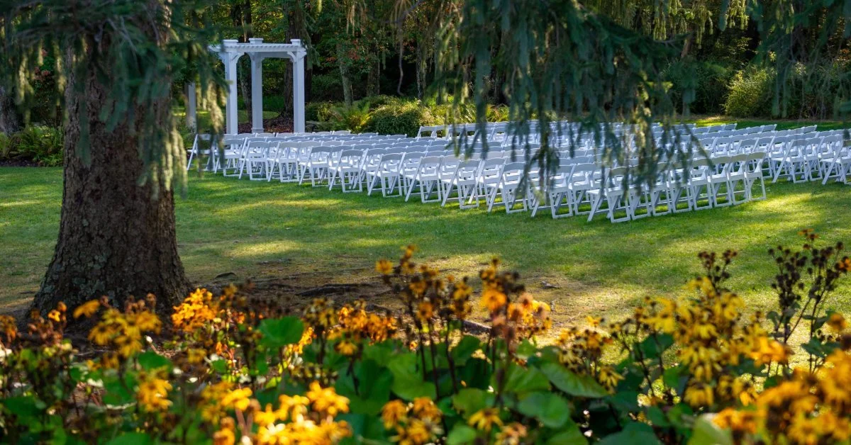 An outdoor wedding ceremony site with rows of white folding chairs arranged on a manicured lawn, facing a white arbor and surrounded by mature trees and yellow garden flowers.