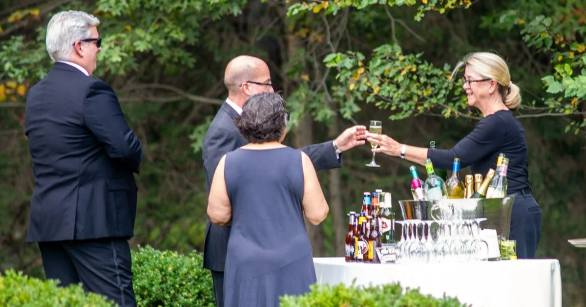 People standing near an outdoor bar table with glasses, bottles, flowers, greenery, and trees in the background.