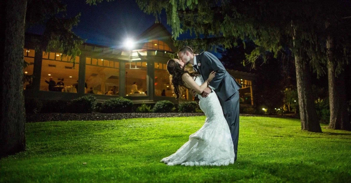 A man and a woman are in the middle of a grassy field. The man is kissing the woman as he dips her down.