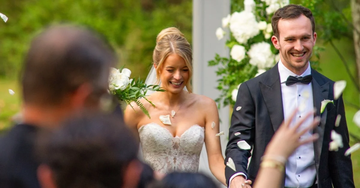 A couple walking outdoors during a wedding ceremony while guests stand nearby and flowers frame the aisle.