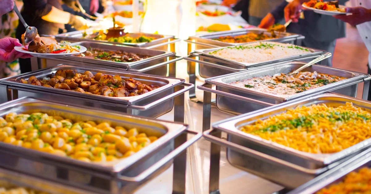 Catered food served buffet style with pans and serving spoons. There are colorful side dishes with meat and vegetables.