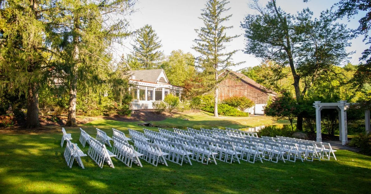 An outdoor wedding ceremony area with rows of white chairs on grass, tall trees, and a white house in the distance.
