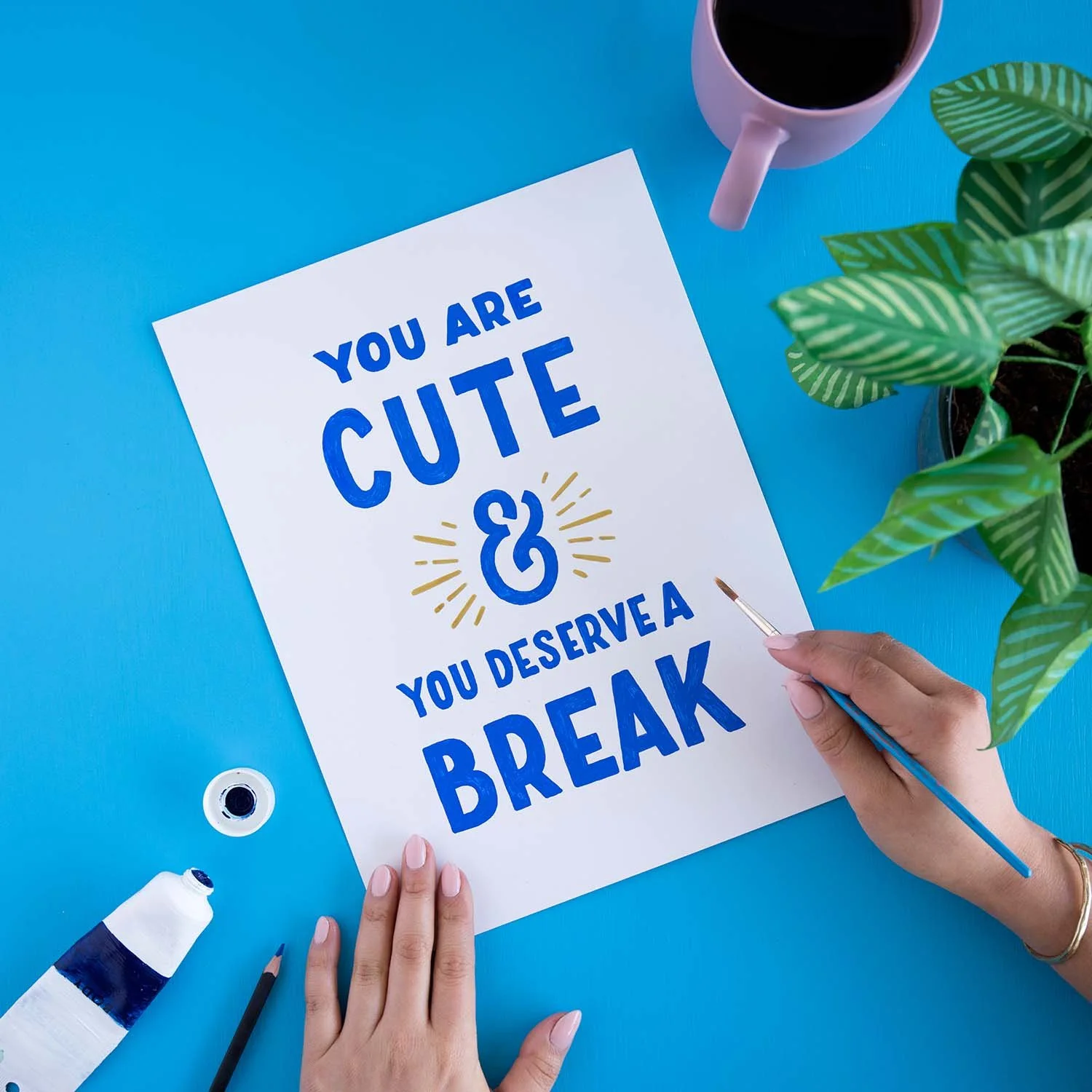 Hands painting 'You are cute and you deserve a break' on paper, surrounded by paint tube, pencil, coffee mug, and paper plant on a medium blue background.