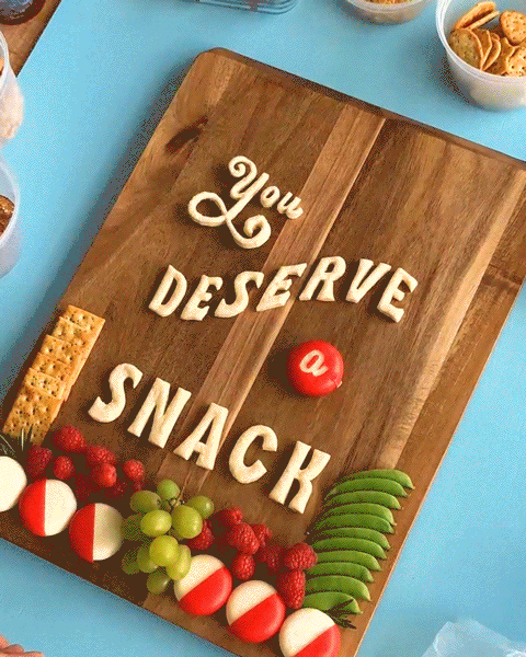 Assembly of the "You Deserve a Snack" charcuterie layout made from Babybel Cheese, crackers, fruit, vegetables, nuts, and herbs.