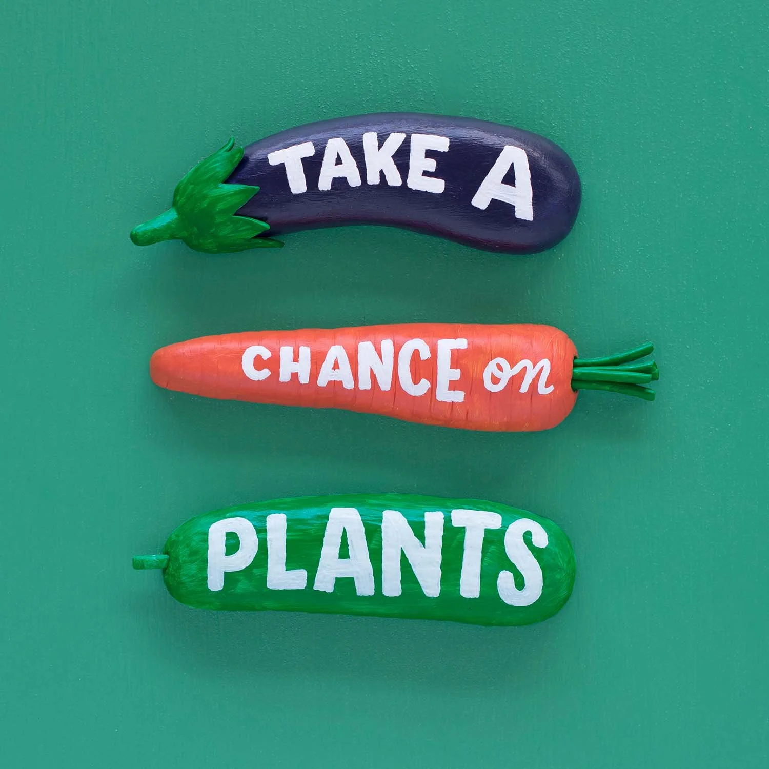 'Take a chance on plants" painted onto an eggplant, carrot, and cucumber resting on a green background.